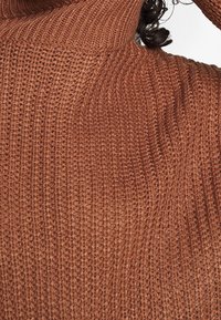 Gros plan sur une personne portant un pull épais en maille de couleur rouille avec un col roulé côtelé haut.