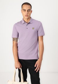 Lavendel polo skjorte med klassisk krave, tre knapper og en liten gul logo på venstre bryst; modell med en beige tote bag.