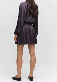 Dunkellila Seidenkleid mit langen, gepufften Ärmeln und einem plissierten Rock. Glatte Textur, tailliert und knielang. Schwarze flache Schuhe.
