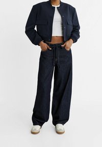 Navy bomber jacket with snap buttons, cropped design, paired with wide-leg dark denim pants and white sneakers with tan soles.