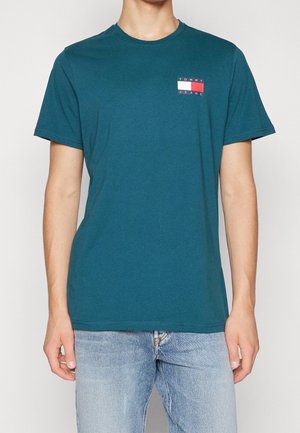 Mand iført en teal Tommy Jeans t-shirt med et lille hvidt og rødt logo på brystet, parret med lyseblå jeans.