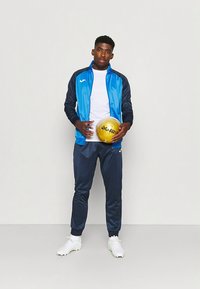 Modra in mornarsko modra športna trenirka z jopico z zadrgo in ujemajočimi hlačami, kombinirana z belimi športnimi čevlji. Drži zlato obarvano žogo.