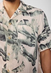 Camisa de manga corta con botones, fondo beige y patrón de hojas tropicales en verde oscuro. Cuenta con un bolsillo en el pecho y una textura sutil.