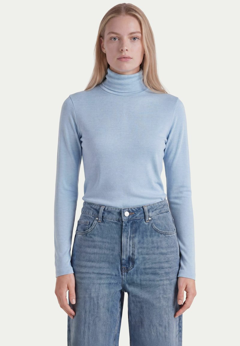 Femme portant un pull à col roulé bleu clair et un jean bleu taille haute, debout devant un fond uni.