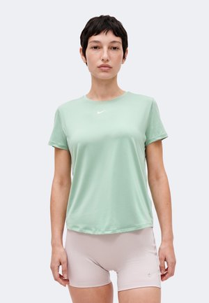 Młoda osoba z krótkimi ciemnymi włosami ma na sobie jasnozieloną koszulkę Nike i beżowe spodenki Nike, stoi na jednolitym jasnym tle.