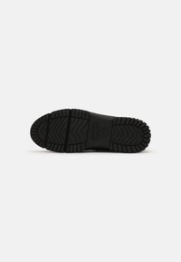 Black rubber shoe sole with deep tread patterns, featuring a curved silhouette, raised logo detail, and a smooth texture.