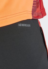 Aeroready sportkleding met een oranje top, zwarte onderkant met logo en gestructureerde rode accenten. Materiaal lijkt lichtgewicht en rekbaar te zijn.