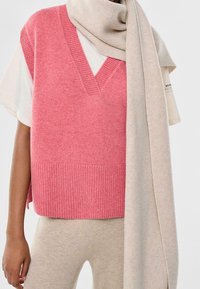 Pull sans manches rose en tricot avec un col en V et un ourlet côtelé, associé à une écharpe beige clair côtelée, drapée lâchement autour du cou.