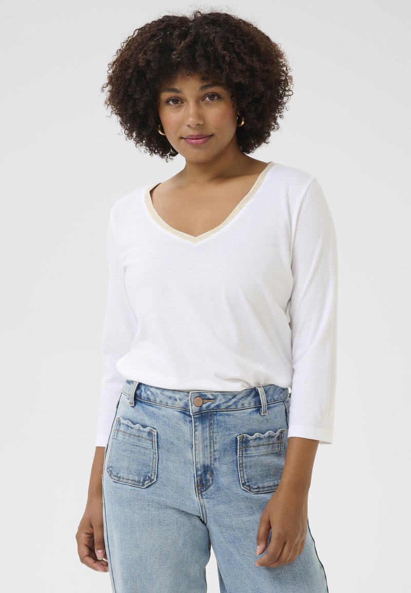 Weißes Langarm-V-Ausschnitt-Shirt mit goldener Verzierung am Ausschnitt, kombiniert mit hochtaillierten, hellblauen Jeans mit gewellten Gesäßtaschen.