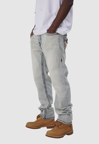 Jasnoniebieskie dżinsy o luźnym kroju, z kontrastowymi szwami i postrzępionymi brzegami, w zestawieniu z brązowymi sznurowanymi butami roboczymi.