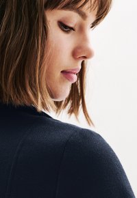 Primo piano di una donna con capelli castani lunghi fino alle spalle e pelle chiara, che indossa un indumento in tessuto di un blu scuro. I suoi tratti del viso sono delicati.