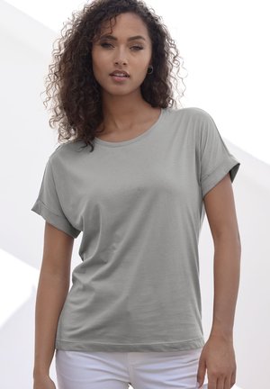 Gray short-sleeve t-shirt in soft fabric with a round neckline and folded sleeves. Worn with white pants, against a bright background.