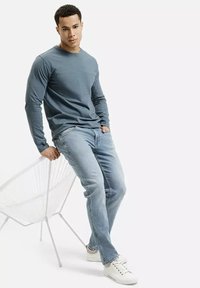 Długosługa, szaro-niebieska koszula bawełniana z okrągłym dekoltem, w połączeniu z jasnoniebieskimi dżinsami i białymi trampkami, oparta o białe krzesło.