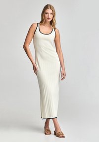 Ribbed, cream-colored maxi dress with black trim on neckline and hem, featuring a fitted silhouette and side slit, worn with brown sandals.