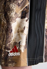 Calções Adidas com um design em camuflagem de tons castanhos e brancos, com painéis laterais pretos, logótipo vermelho e tecido texturizado.