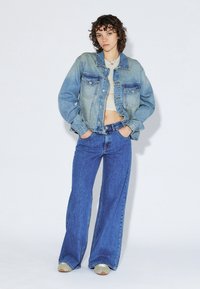 Denim jacket in light blue, with two chest pockets and button closure. Wide-leg blue jeans, textured finish, complemented by beige sneakers.