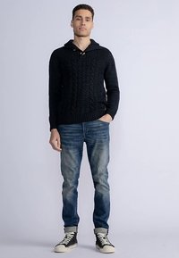 Schwarzer gestrickter Pullover mit Zopfdesign und Kapuze. Kombiniert mit verwaschenen blauen Jeans und High-Top-Sneakers. Modell steht mit Blick nach vorne.