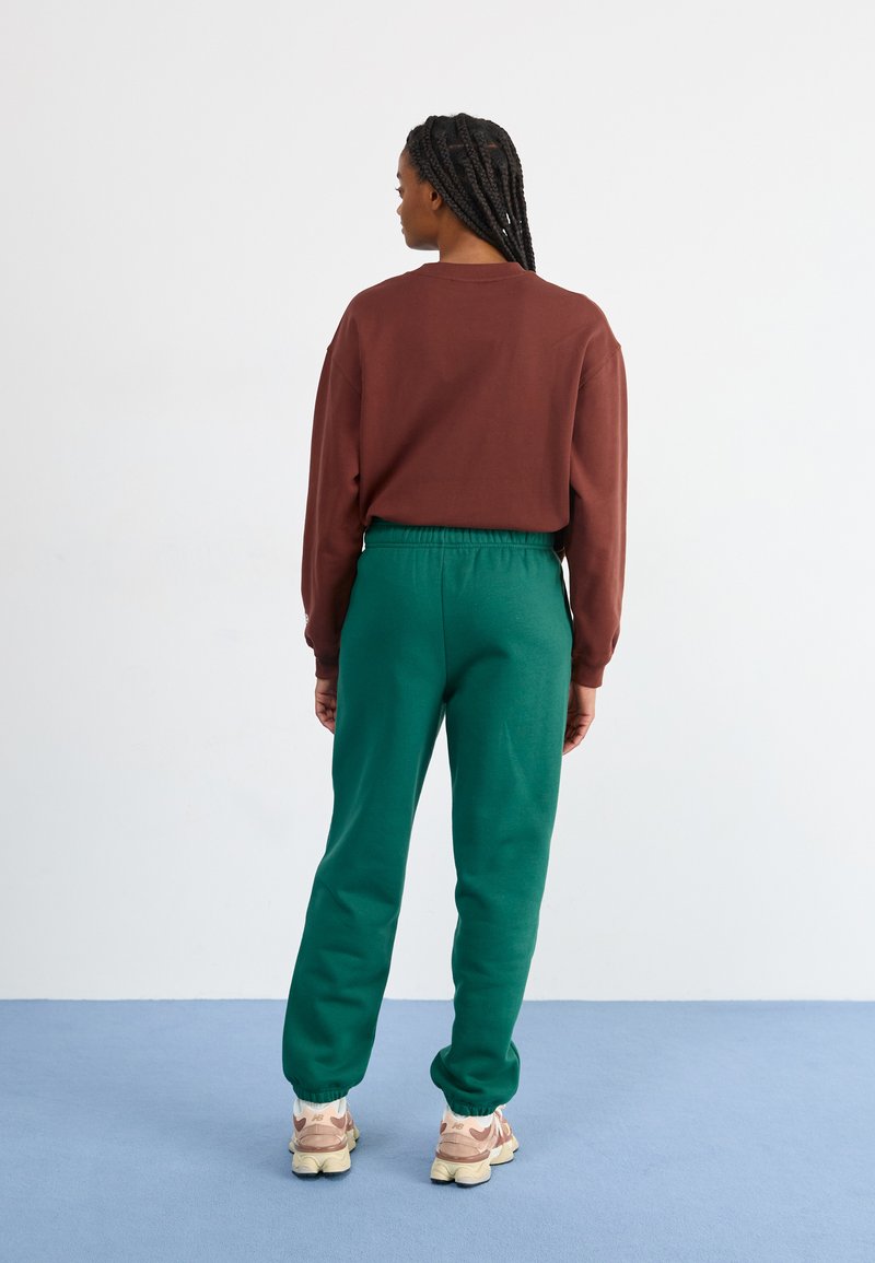 Bruine lange-mouwen top. Groene jogger broek met elastische taille en boorden. Lichtgekleurde sneakers aan de voeten. Eenvoudige, casual outfit.
