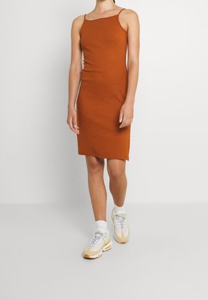 Personne portant une robe sans manches couleur rouille avec des bretelles fines et des baskets blanches, marchant devant un fond clair uni.