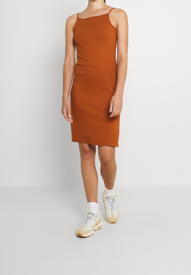 Personne portant une robe sans manches couleur rouille avec des bretelles fines et des baskets blanches, marchant devant un fond clair uni.