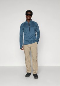 Blå fleecetröja med dragkedja och ett texturerat mönster, kombinerad med beige cargobyxor. Modellen bär svarta solglasögon och svarta skor.