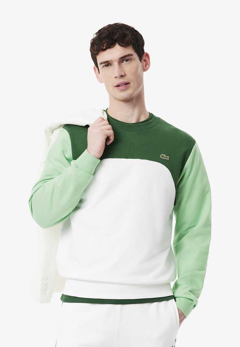 Sudadera en bloque de color verde y blanco con cuello redondo y puños acanalados. Presenta el logo de Lacoste en el pecho y una textura suave.