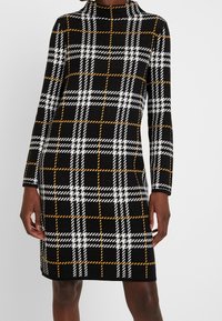 Femme portant une robe noire mi-longue avec des motifs à carreaux blancs et orange et des manches longues, debout devant un fond uni.