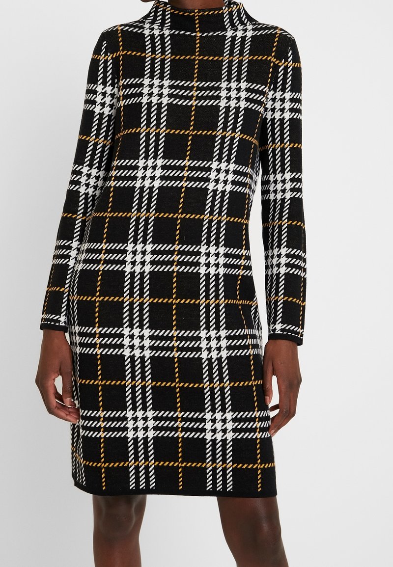 Femme portant une robe noire mi-longue avec des motifs à carreaux blancs et orange et des manches longues, debout devant un fond uni.