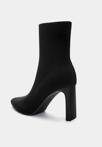 Bottine noire en tricot avec un bout pointu et un talon aiguilé haut. Présente un bord supérieur côtelé et une texture lisse sur le corps principal.