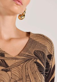 Bruine gebreide trui met zwart bladerenpatroon, voorzien van een V-hals. Gouden statementoorbel met ronde vormen en glanzende textuur.