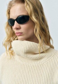 Maglione beige a coste con un alto colletto a pieghe. Occhiali da sole ovali neri. Lunghi capelli biondi e ondulati che incorniciano il viso. Sfondo semplice.