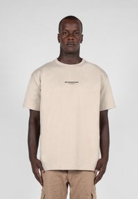 Beige bomulds t-shirt med rund hals, korte ærmer og teksten "METAMORPHOSIS" i sort på forsiden. Båret med khaki cargo bukser.