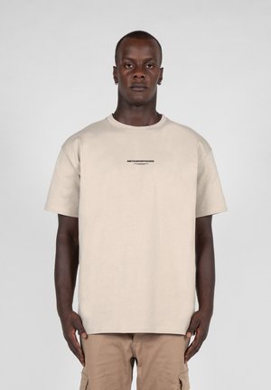 Beige bomulds t-shirt med rund hals, korte ærmer og teksten "METAMORPHOSIS" i sort på forsiden. Båret med khaki cargo bukser.