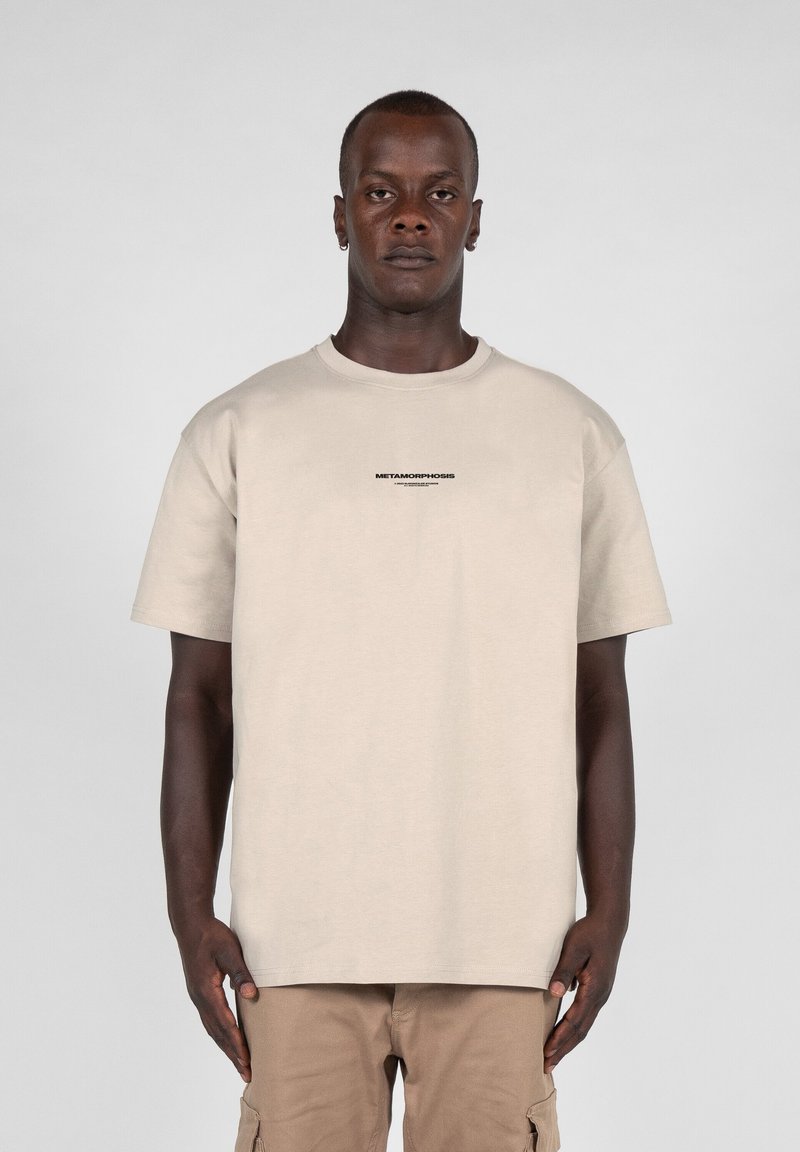Beige bomulds t-shirt med rund hals, korte ærmer og teksten "METAMORPHOSIS" i sort på forsiden. Båret med khaki cargo bukser.