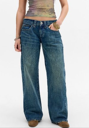 Personne portant un jean bleu ample, un haut court tie-dye, des bracelets et des chaussures marron, debout avec une main dans la poche.