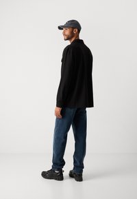 Mand iført sort jakke, blå jeans, sorte sneakers og en grå baseballcap, der står sidelæns mod en hvid baggrund.