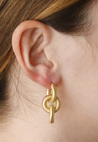 Pendientes de metal dorado en un diseño entrelazado que presenta formas suaves y redondeadas con distintos grosores, colgando de un gancho simple.