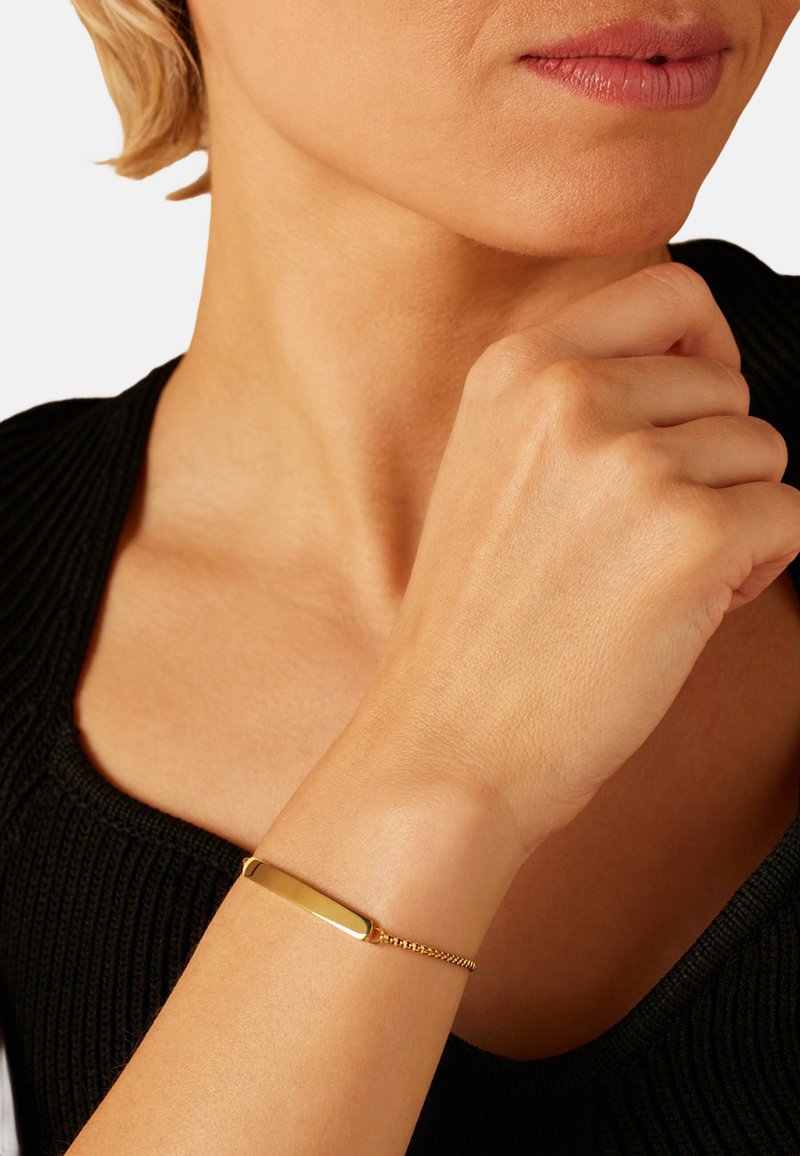 Gouden armband met een slanke rechthoekige plaat en een schakeldesign, gedragen om een pols met een warme huidskleur, tegen een zwart geribbeld topje.