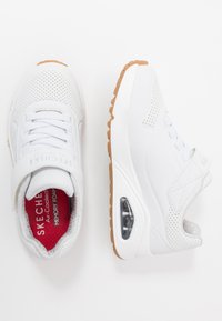Witte Skechers sneakers met een geperforeerde bovenkant, verstelbare band en een tan kleurige rubberen zool met zichtbare luchtkussen. Rode gevoerde binnenzool.