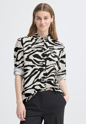 Woman wearing a black-and-white zebra print button-up shirt with rolled sleeves and black pants, standing with one hand in pocket.