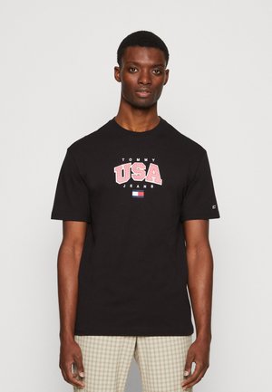 Sort bomulds T-shirt med en stor "TOMMY USA" grafik i rød og hvid, rund halsudskæring og korte ærmer. Båret med lyse ternede bukser.