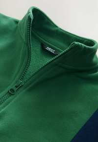Grøn fleecepullover med høj krave, halv-zip foran og kontrastfarvet navy sidepanel. Blød tekstur med en glat finish. Brandlabel synlig.
