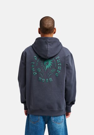 Osoba w ciemnoszarej bluzie z kapturem z zielonym motywem słońca yin-yang i dłoni oraz tekstem na plecach, w połączeniu z niebieskimi dżinsami, stojąca tyłem.