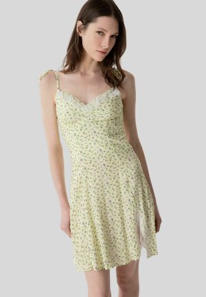 Donna che indossa un vestito sottoveste di colore giallo pallido con stampa floreale, spalline sottili e rifiniture in pizzo sullo scollo e sull'orlo.