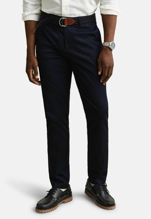 Hombre vistiendo pantalones azul marino oscuro con cinturón de cuero negro, camisa blanca con las mangas remangadas, zapatos negros con suelas marrones y reloj de pulsera plateado.