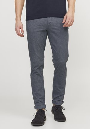 Homme portant un pantalon slim gris foncé avec des revers et des chaussures à lacets noires, debout devant un fond clair uni.