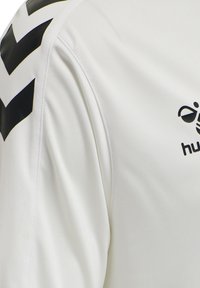 Manche de maillot de sport blanche avec des bandes en chevron noires sur l'épaule et logo partiel noir de la marque sur la poitrine.