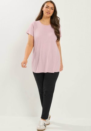 Vrouw met lang bruin haar, gekleed in een losse lichtroze T-shirt, zwarte broek en witte sneakers met zwarte strepen, staand tegen een effen achtergrond.