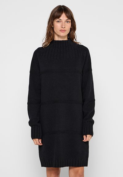 Femme aux cheveux bruns portant une robe pull à col roulé noire longue, debout devant un fond uni gris clair.