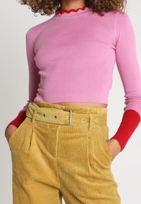 Eng anliegendes rosa geripptes Langarmshirt mit rotem, wellenförmigem Kragen, kombiniert mit hoch taillierten gelben Cordhosen mit Gürtelschlaufen und Taschen.